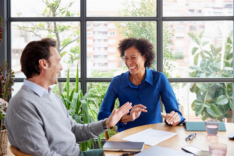 Deux professionnels en pleine réunion sourient et échangent dans un bureau moderne et lumineux entouré de plantes, illustrant une ambiance de travail positive et collaborative