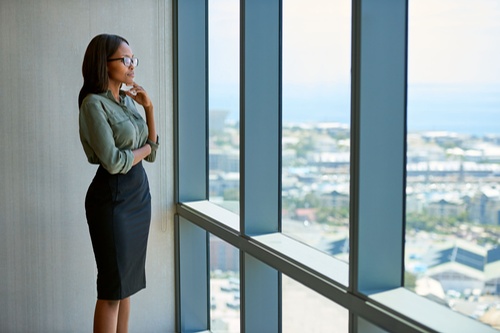 Une jeune femme d'affaires séduisante portant des lunettes regarde pensivement à travers les fenêtres du bureau l'horizon de la ville